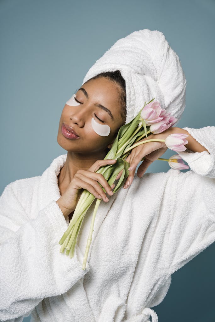 Tranquil African American female with eye patches in white bathrobe and towel on head standing with closed eyes and bouquet of tulips against blue background